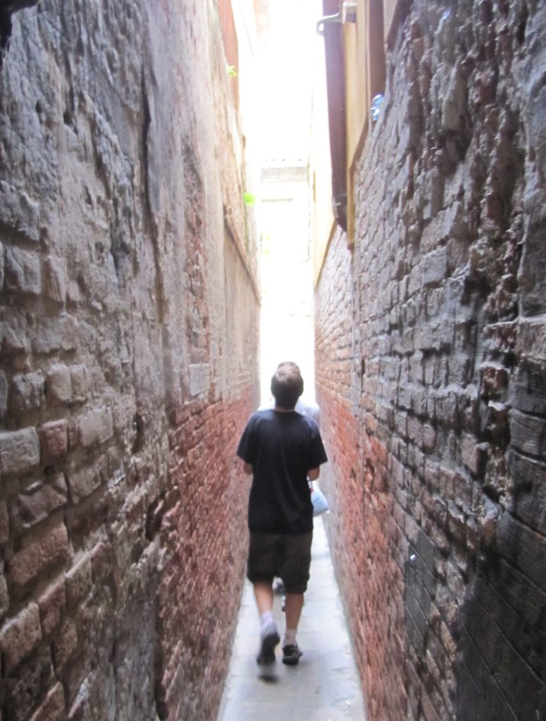Venice with teens: boy on narrow path between high brick walls. Copyright©2015 mapandfamily.com 
