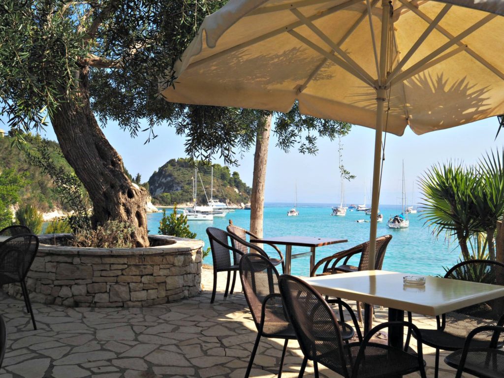 Taverna terrace with umbrella and large olive tree overlooking bay and Harami beach. Copyright © 2017 mapandfamily.com 