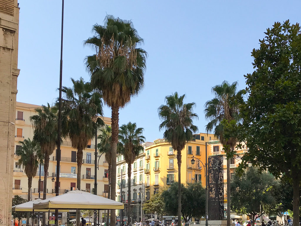 A square in Naples with pastel coloured buildings and rows of palm trees. 