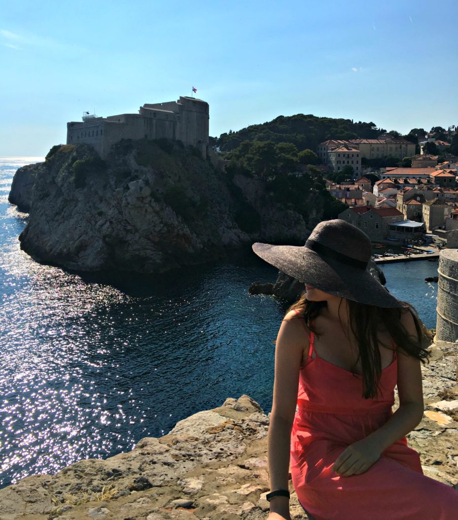 Dubrovnik things to do. Girl in a red dress and black sun hat sitting on the walls by the sea. Copyright©2017 mapandfamily.com