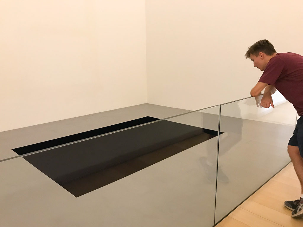 Boy looking over glass barrier at dark rectangle artwork on floor of museum