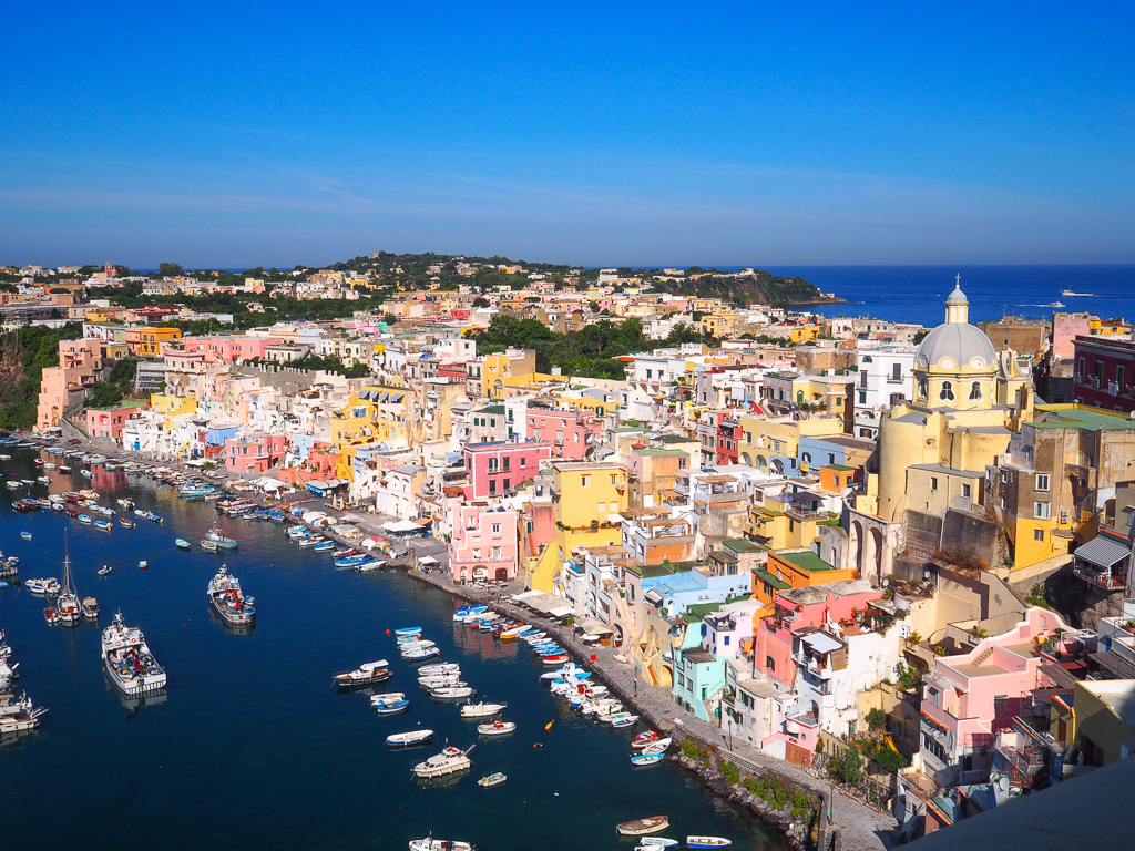 Where is hot in Europe in September? Colourful Corricella on Procida island in the Bay of Naples Italy. Pastel-coloured houses overlook dark blue harbour. Copyright@2019 mapandfamily.com 