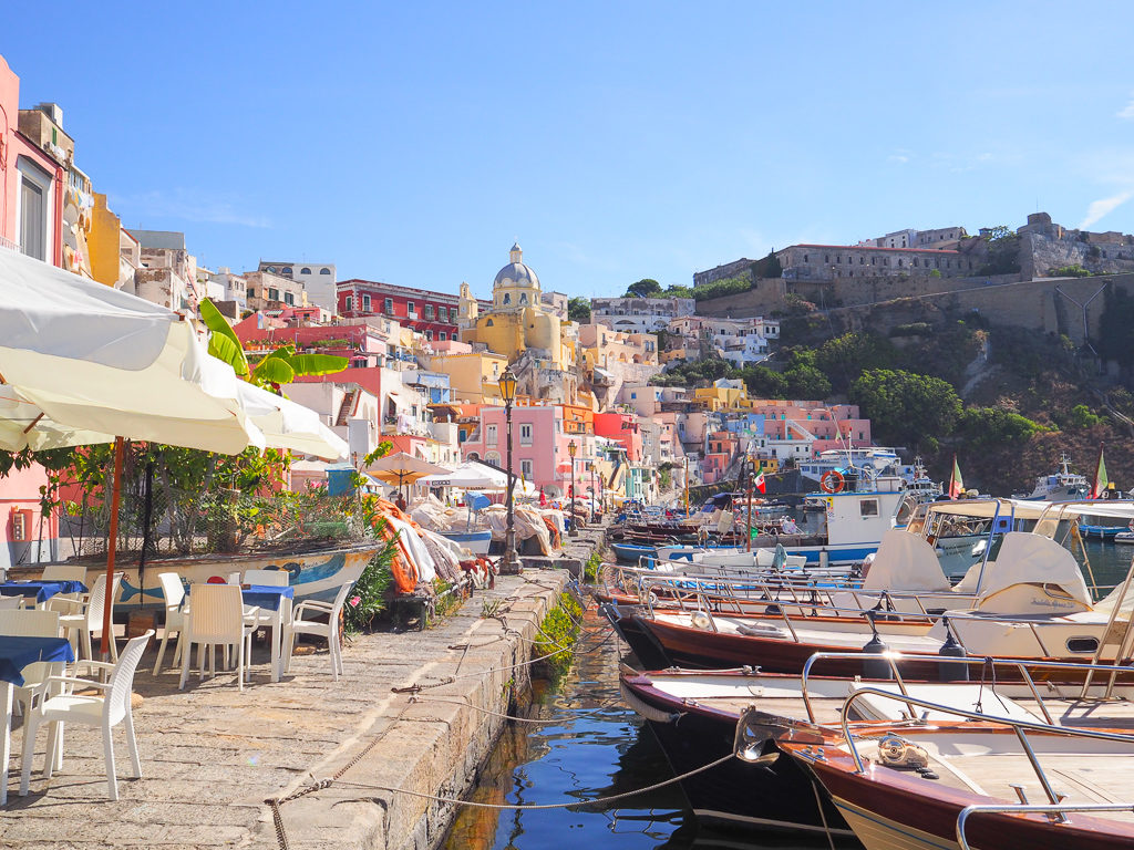 On the quayside in Corricella, Procida island with umbrellas, chairs and boats moored up. Copyright©2019 mapandfamily.com 