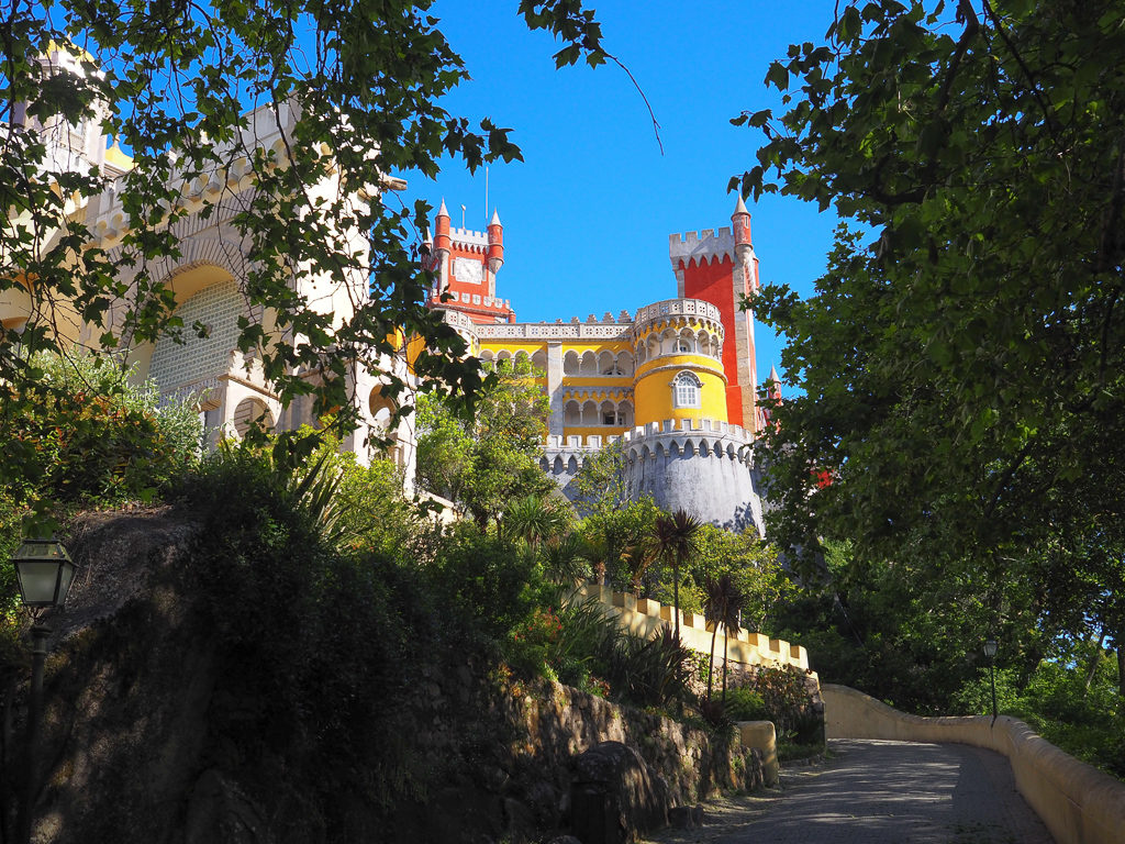 Yellow and red castellated towers of Pena Palace through trees. Copyright ©2019 mapandfamily.com 