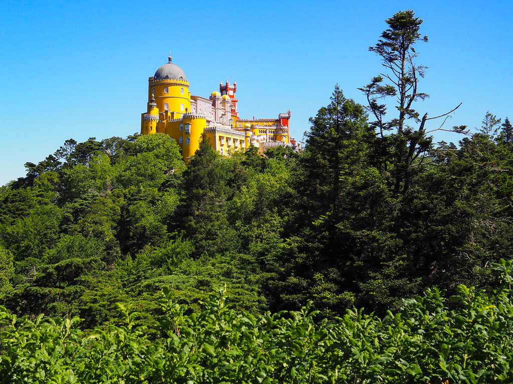 Brightly hued Pena Palace in shades of yellow and red on a green hillside. Copyright@2025 NancyRoberts