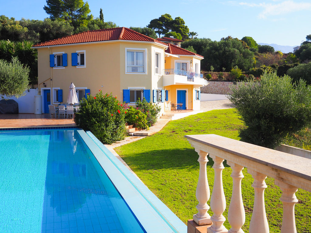 Kefalonia villa, large sand coloured house with balcony, blue shutters and  pool. Copyright ©2019 mapandfamily.com 