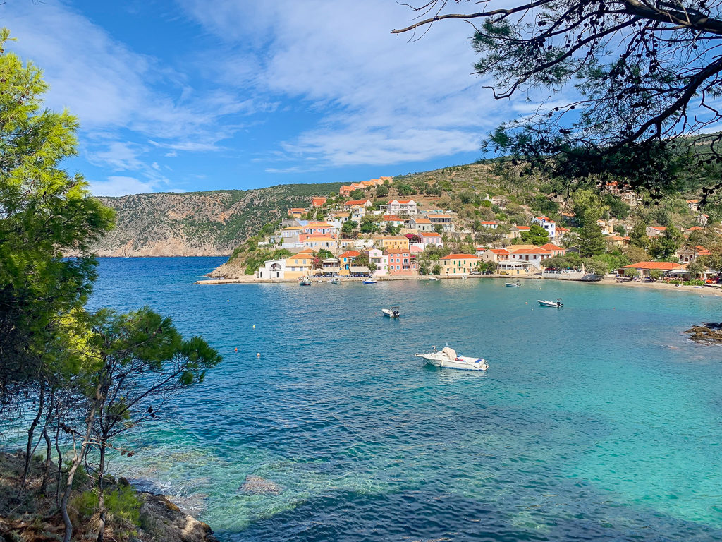Assos Kefalonia. View of bay with trees, boats, blue sea and colourful houses. Copyright ©2019 mapandfamily.com 