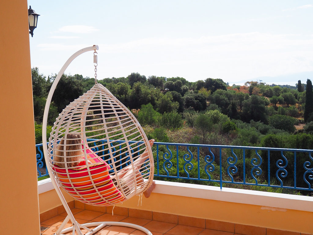 Backview of person sitting in white pod shaped swing seat on balcony with orange cushions facing a rural view to the sea. Copyright ©2019 mapanfamily.com 