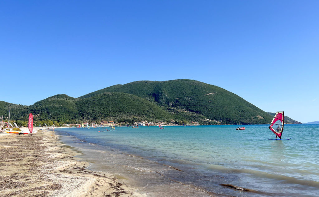 Vasiliki beach, Lefkada. Red windsurf in foreground, Sand and shallow water with green hills behind. Copyright@ 2022 mapandfamily.com 