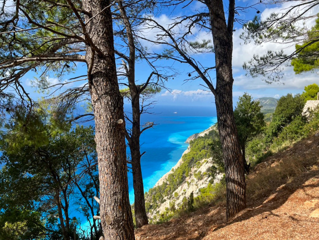 View through pine trees of cliffside and dazzling turquoise and indigo sea in the distance. Copyright@ 2022 mapandfamily.com 