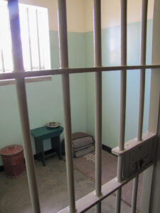 Small prison cell with folded blanket, stool and bars on door and window. Copyright@2023 mapandfamily.com