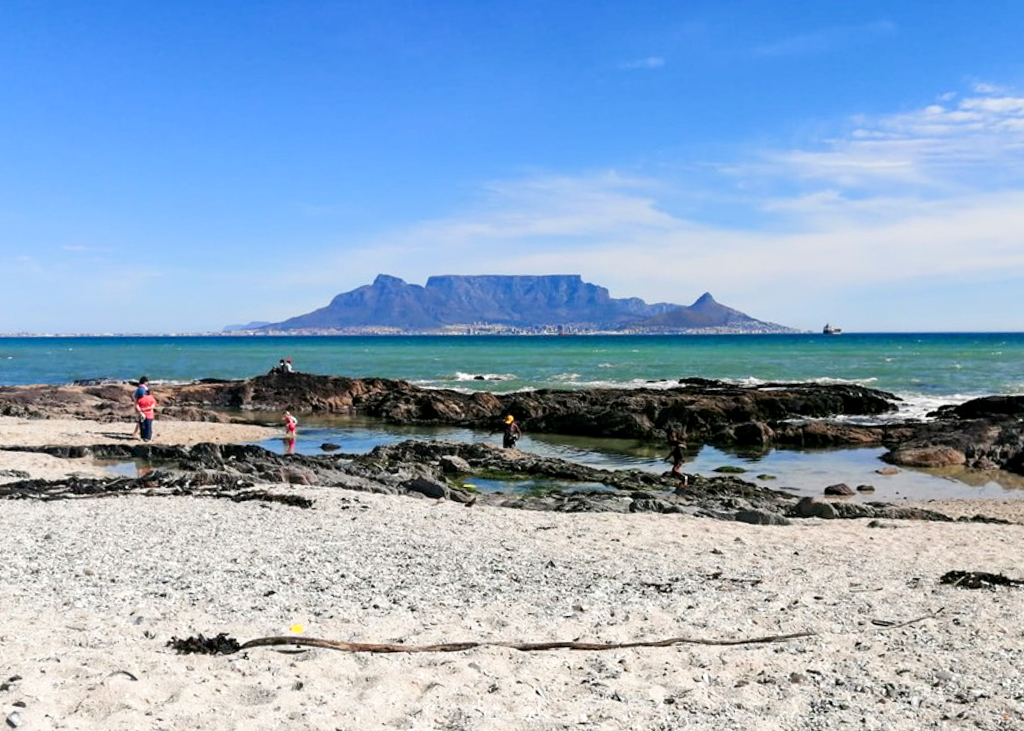 People paddling in a large rock pool on a beach with the outline of Table Mountain on the other side of the bay. Copyright@2023 reserved to photographer via mapandfamily.com