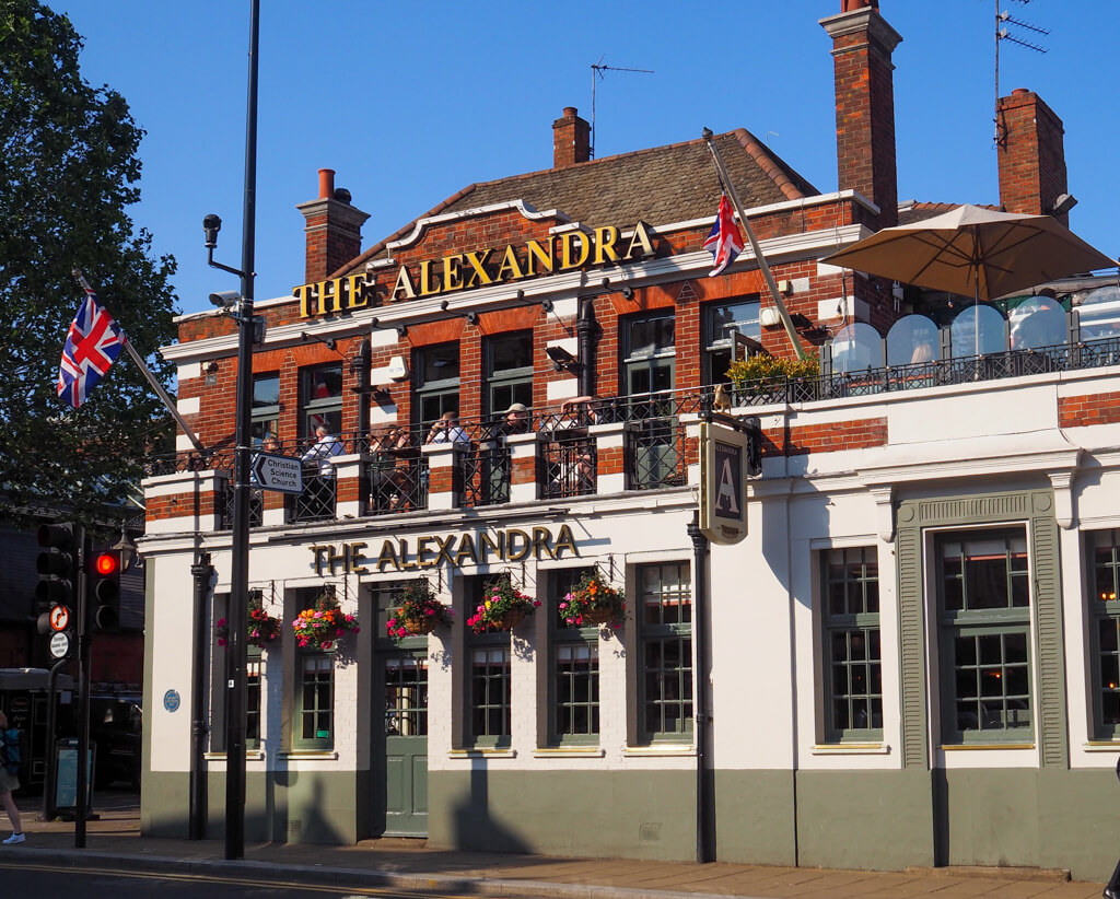 Live in Wimbledon: exterior of pub with first floor terrace and name The Alexandra on wall above hanging baskets of flowers. Copyright@2023mapandfamily.com