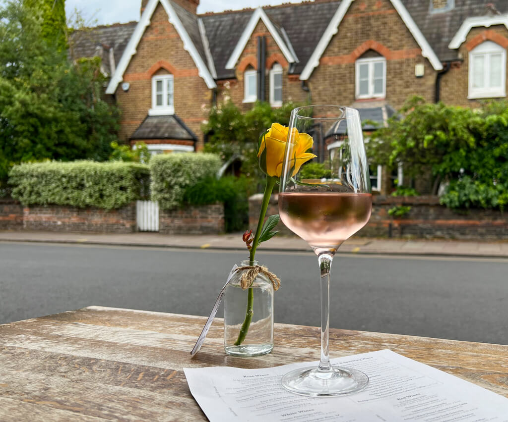 A glass of rose wine and a yellow rose in a tiny vase on a pavement side table top with a road and cottages in background. Copyright@2023mapandfamily.com 