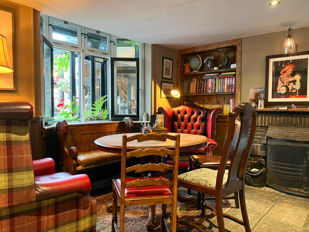 Round table with collection of varied vintage dining chairs in cosy corner of pub with bookshelves and fireplace. Copyright@2023mapandfamily.com 