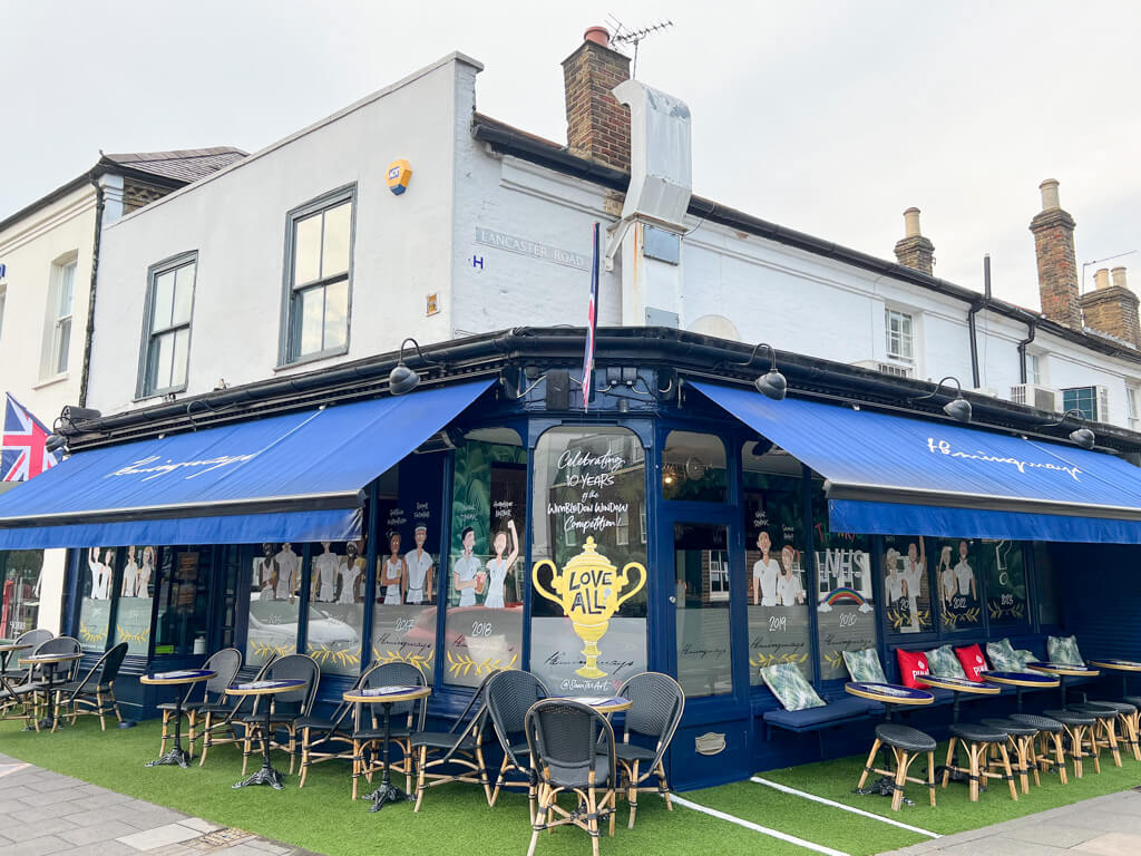Live in Wimbledon: Hemingways bar with blue awnings, tables and chairs outside and cartoons of tennis players drawn onto the windows. Copyright@2023mapandfamily.com