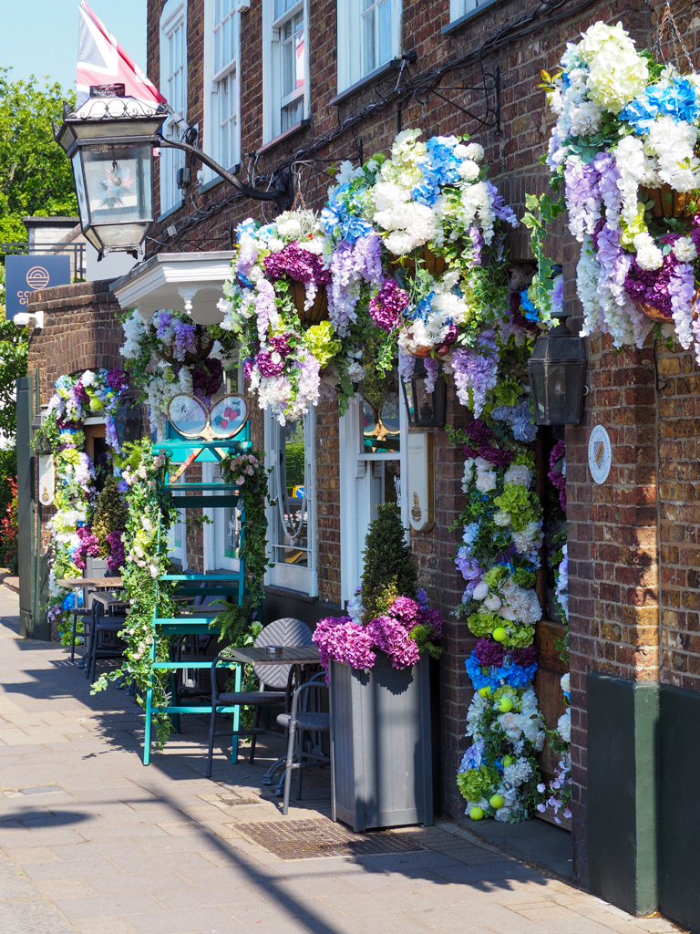 Exterior of pub with old lantern and hanging baskets packed with flowers in white, purple and blue. An umpire's chair stands outside. Copyright@2023mapandfamily.com 