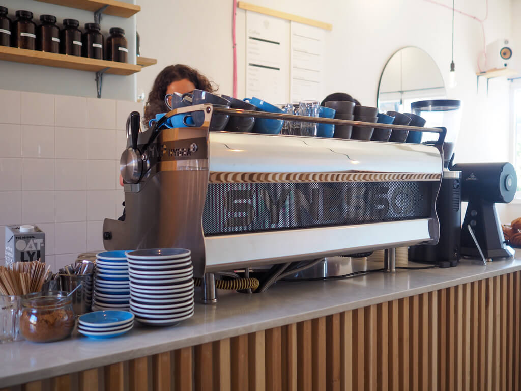 Interior of Wimbledon cafe showing large coffee machine with stacks of blue and grey cups and saucers. Copyright@2023 mapandfamily.com 