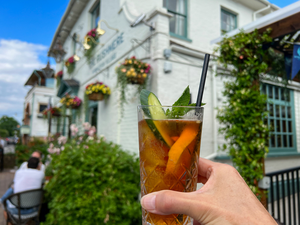 Hand holding up a glass of Pimm's with straw and fruit garnish. Pub in background. Copyright@2023mapandfamily.com 