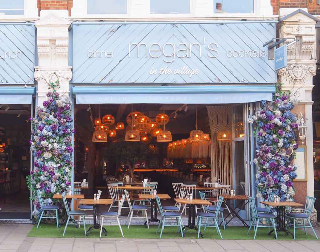 Megan's Cafe which opens onto the pavement with tables and chairs outside, lots of lights with woven shades.  Plus pink, purple and blue flower display. Copyright@2023 mapandfamily.com 