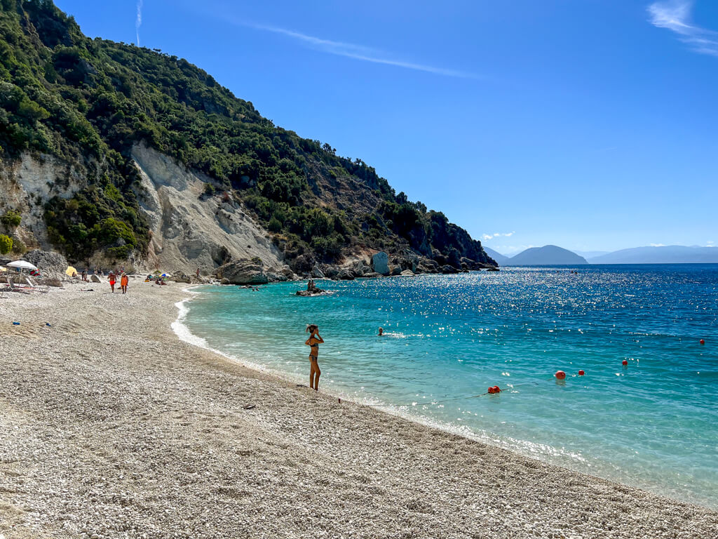 White pebble beach and turquoise sea with green cliffs behind at Agiofili Lefkada beach. Copyright@2023mapandfamily.com 