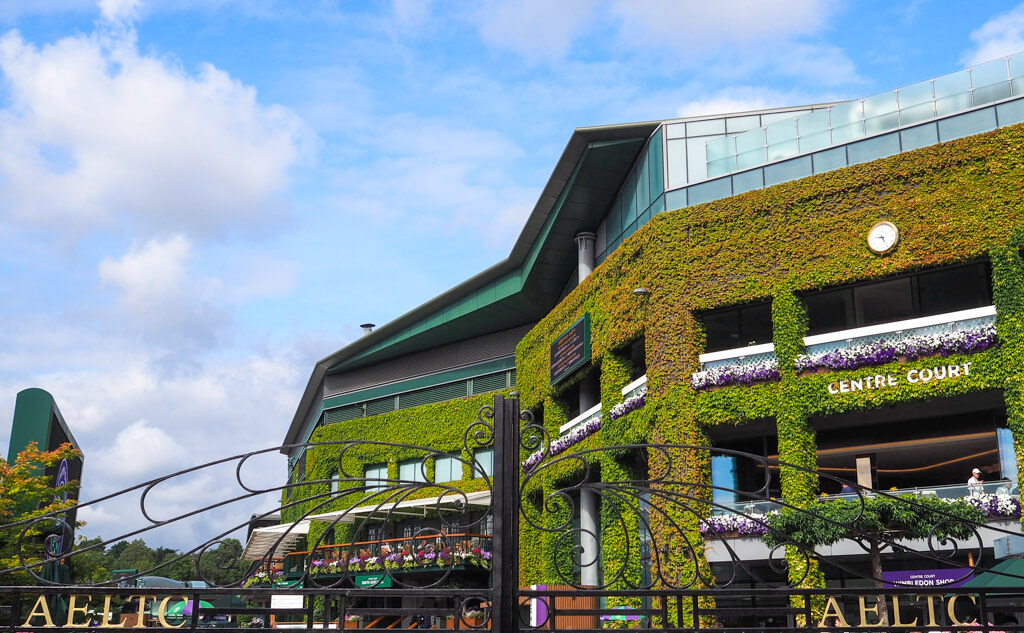 One of the best things to do in Wimbledon London is go to the tennis. View of exterior of Centre Court with green creeper on walls, and gates of All England Club. Copyright@2023mapandfamily.com 