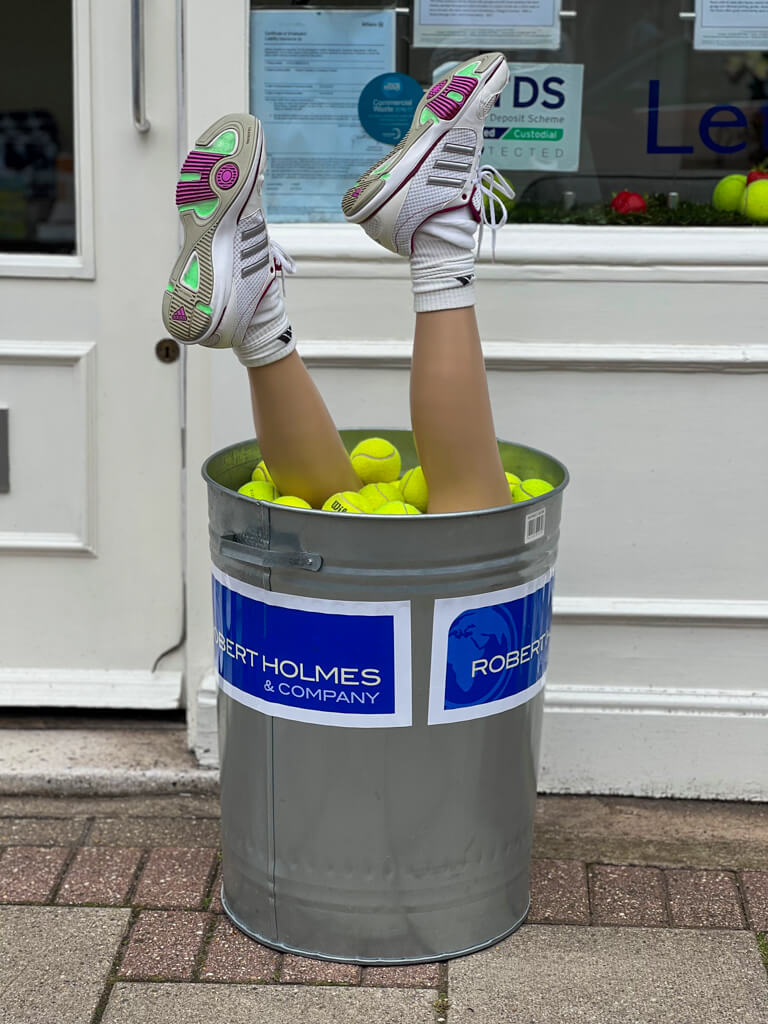 Bucket of balls with mannequin's legs wearing tennis shoes sticking out of it! Tennis themed display at estate agency in Wimbledon. .Copyright@2023mapandfamily.com 
