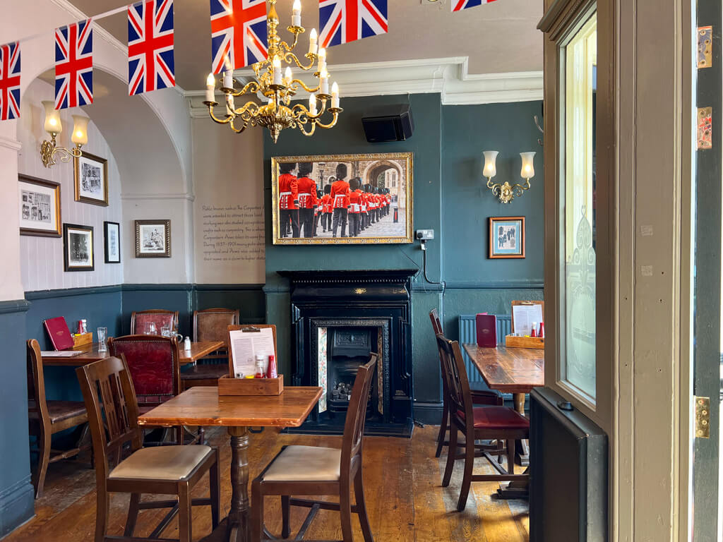 Interior of pub with a large photo of marching Guards on wall above fireplace. Wooden tables and chairs and union flags. Copyright@2023 mapandfamily.com 