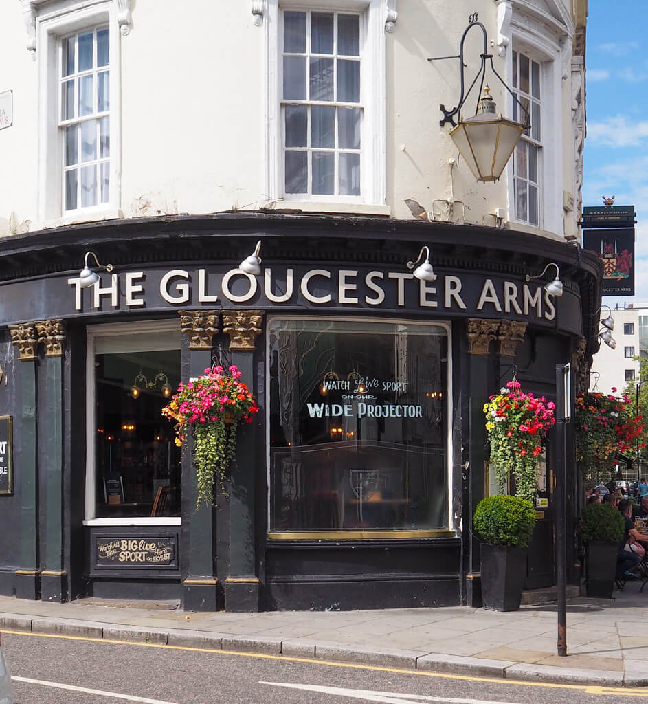 The Gloucester Arms has a black painted exterior with hanging baskets of flowers and a lantern over the door. Copyright@2023 mapandfamily.com