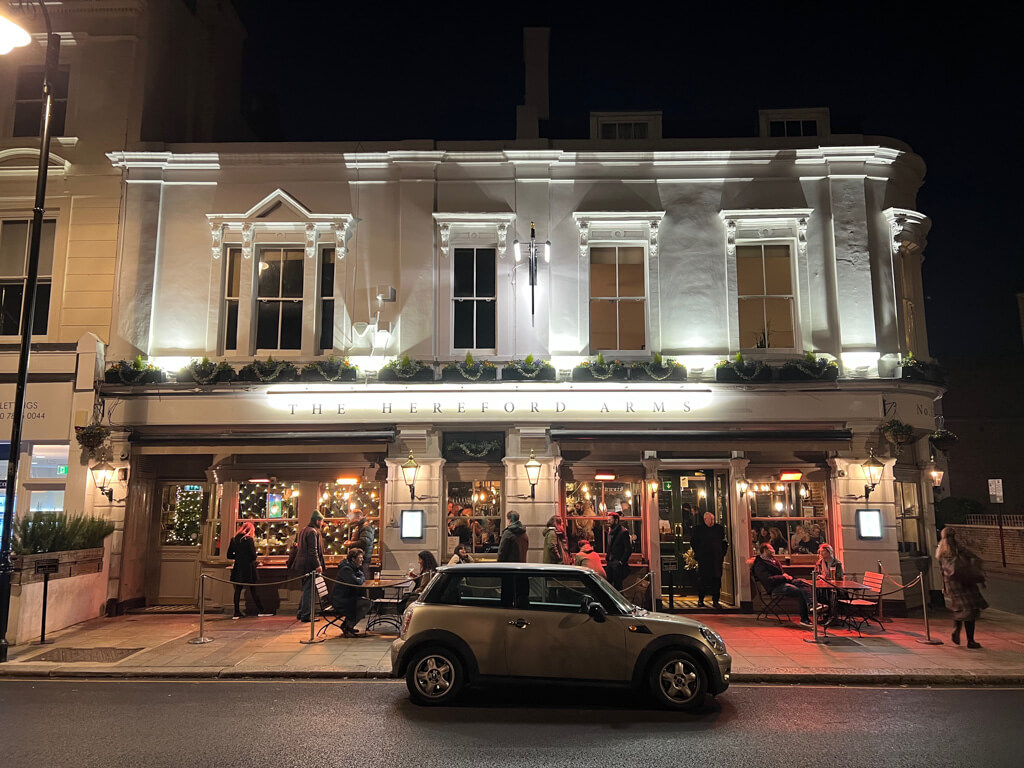 The Hereford Arms in the evening with a few people sitting at the outdoor tables and a cosy glow of light reflected on the pavement. Copyright@2023 mapandfamily.com