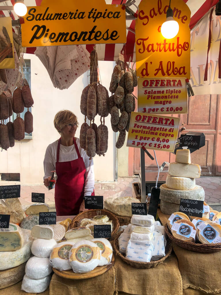 Things to do in Piedmont: visiting a food market: a stall with lady in red apron behind counter with cheese and cured meats. Copyright@2023 reserved to the photographer via mapandfamily.com