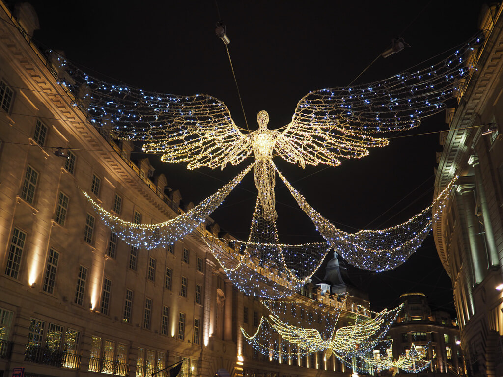 A golden winged angel installation, twinkling with lights above Regent Street with grand terraced buildings on either side. Copyright @2023 mapandfamily.com 