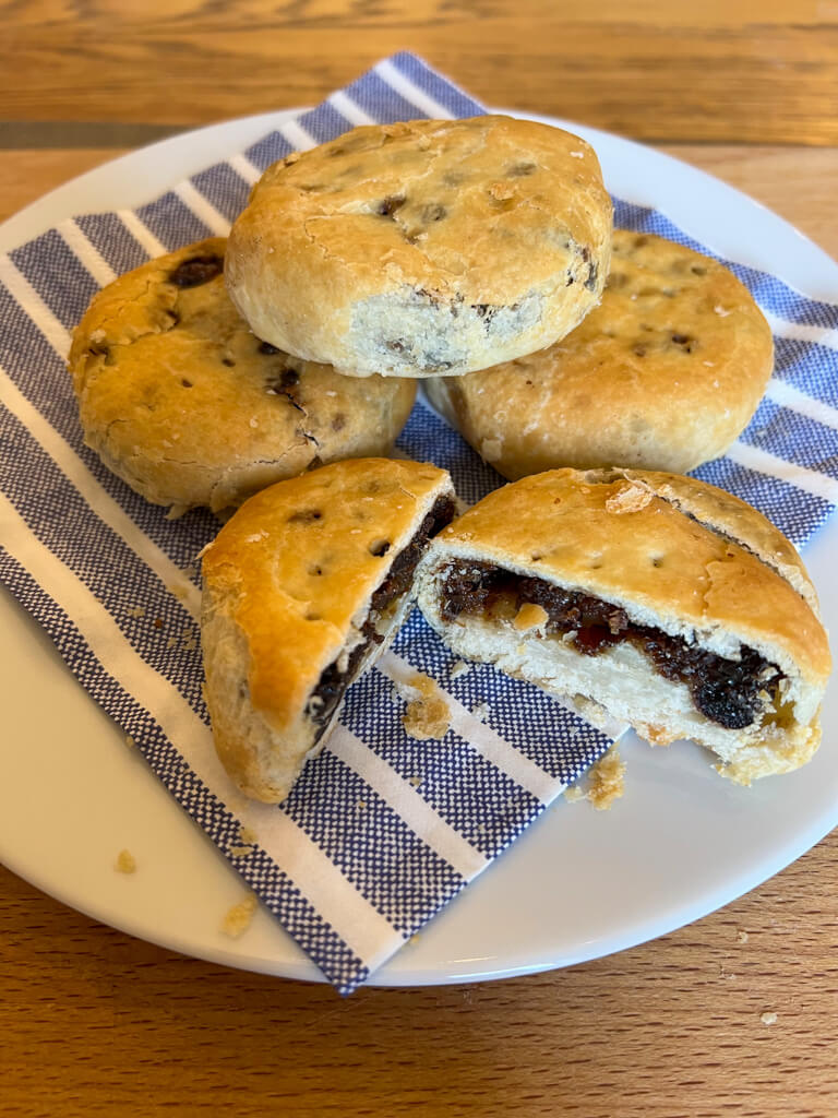 Three Eccles cakes piled on a plate with a blue striped serviette. Plus a bun which has been cut in half. Copyright @2023mapandfamily.com 