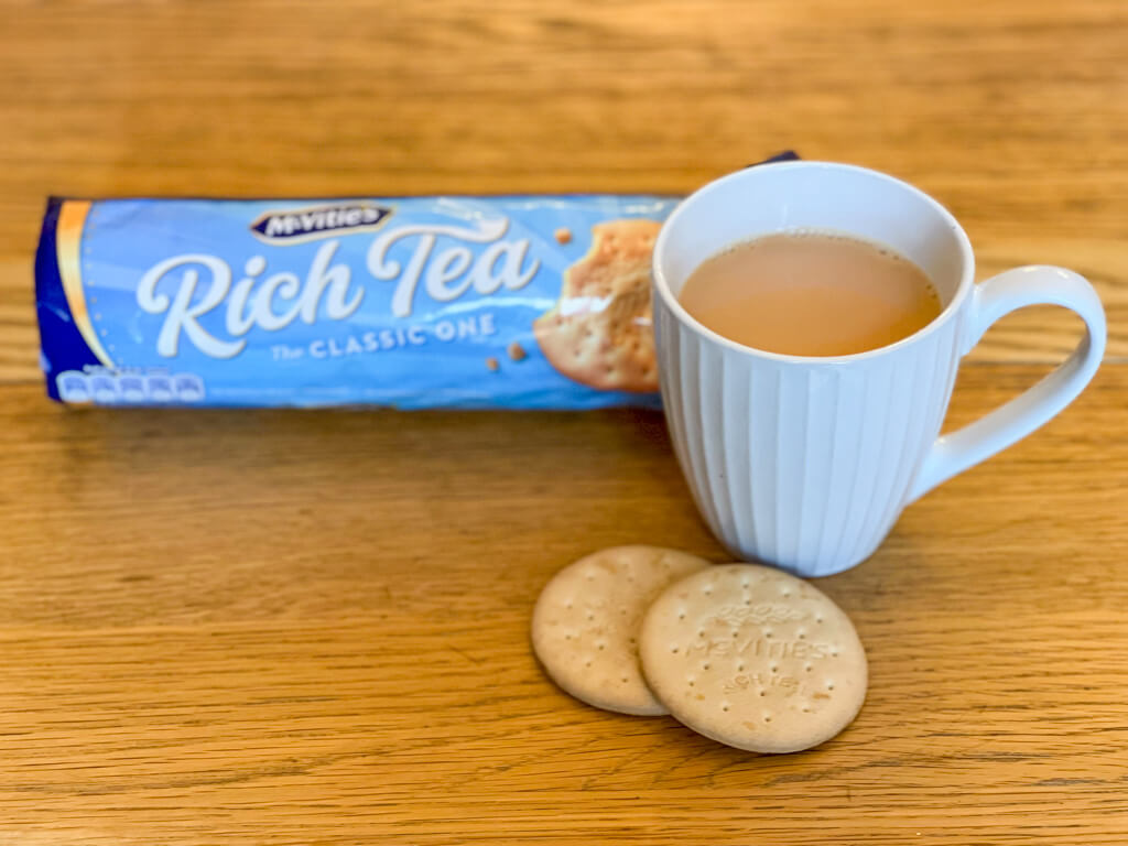 A cup of tea in a white fluted mug with two Rich Tea biscuits and a packet in the background. Copyright 2023@mapandfamily.com