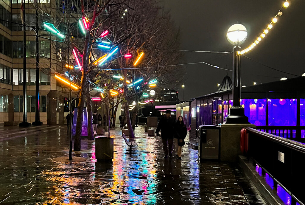 Neon light displays reflects colourfully on wet pavements at Christmas market at night. Copyright@2023 mapandfamily.com 