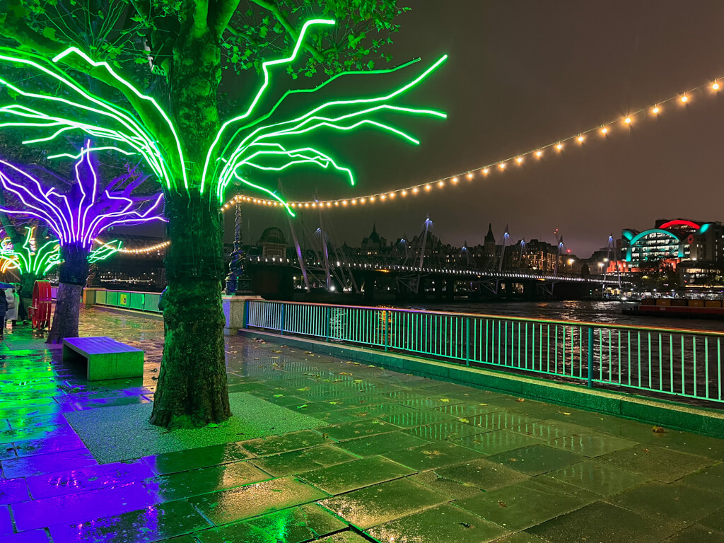 Neon coloured lights in trees on walkway beside river Thames at night. Copyright@2023 mapandfamily.com