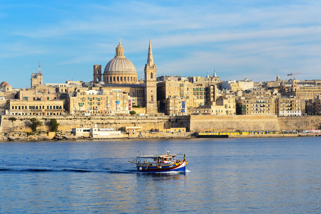 A hot destination in Europe in April is Malta. View of the historic golden city overlooking the sea with a traditional boat in foreground. 