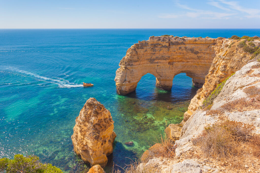 Translucent blue sea and a small boat beside golden rocks and natural arches on the coastline of the Algarve. 