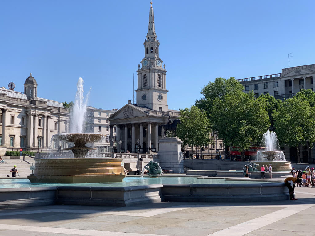A sunny day in Trafalgar Square with fountain in foreground and church steeple backed by blue sky. Copyright@2024NancyRoberts