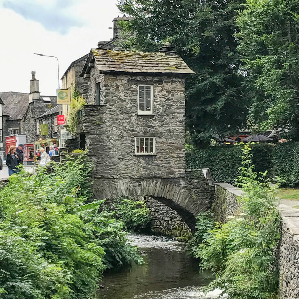 A tiny grey stone building perched on an arched bridge above a stream in the Lake District. Copyright@2025NancyRoberts