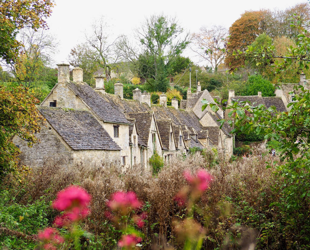 A pretty English countryside scene of  a row of golden stone cottages with pink flowers in the foreground. Copyright@2025NancyRoberts