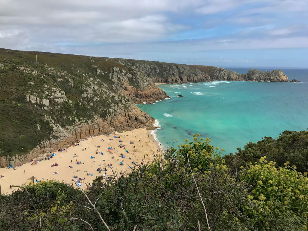 Porthcurno Cornwall, looking down from a cliffside onto a golden sandy beach with turquoise sea.Copyright@2025NancyRoberts