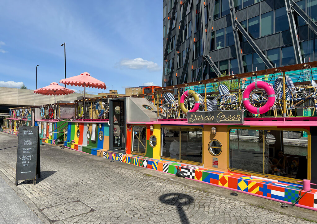 A colourful narrowboat cafe in Paddington with a pink umbrella on upper deck terrace. Copyright@2024 mapandfamily.com