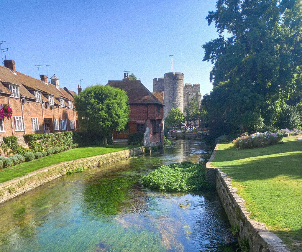 A river running between lawns in a park in Canterbury with houses on one side overlooking the water and a medieval tower ahead. Copyright@2024 mapandfamily.com 