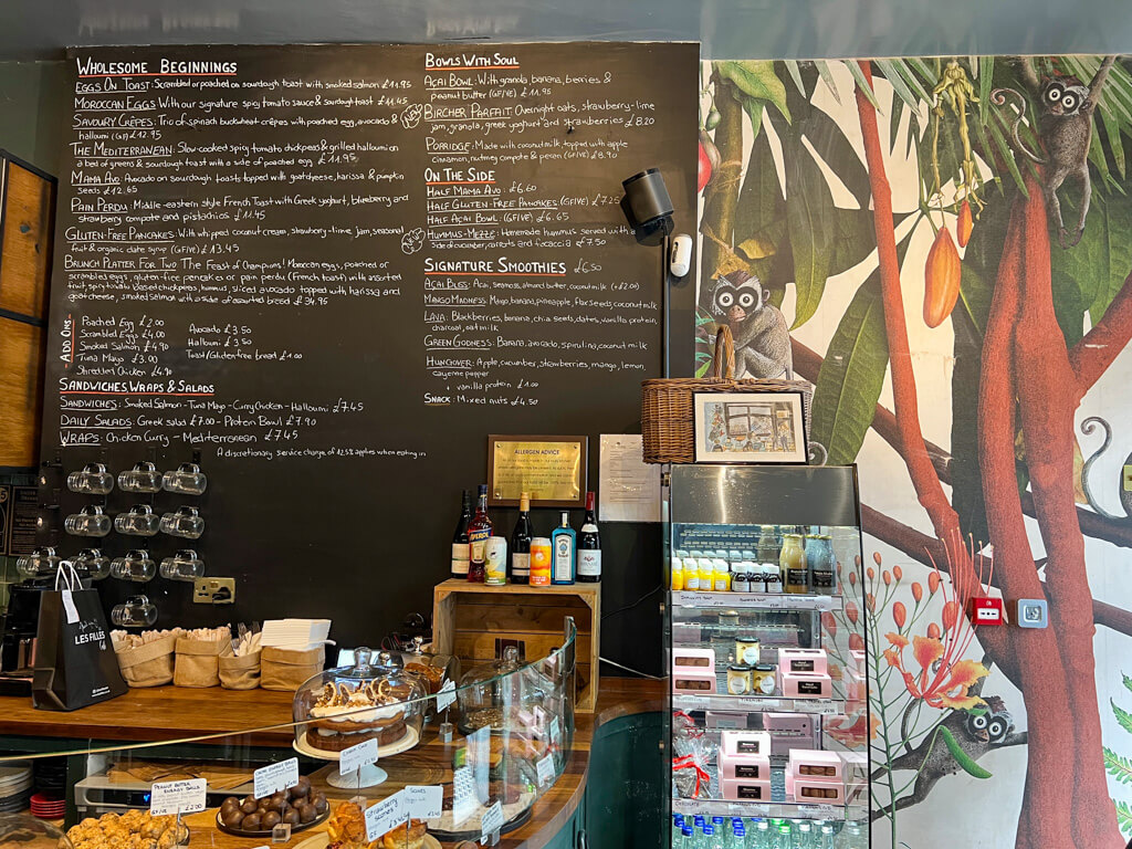 Interior of cafe Les Filles showing a chalkboard menu, a display of cakes and a jungly mural on the wall. Copyright@2024 mapandfamily.com