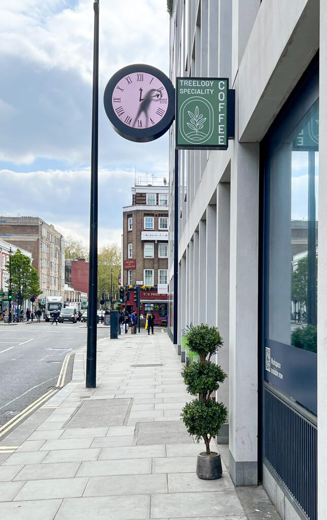 Paddington cafe: exterior of Treelogy with a topiary plant and the wall-hung Real Time clock with a moving figure in the clock face. Copyright@2024 mapandfamily.com