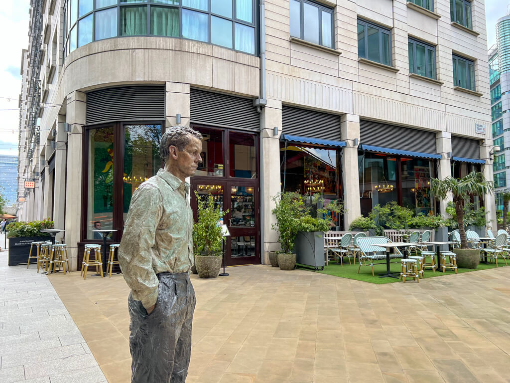 Life-sized sculpture of a man standing outside a cafe with turquoise wicker seating on the terrace. Copyright@2024 mapandfamily.com