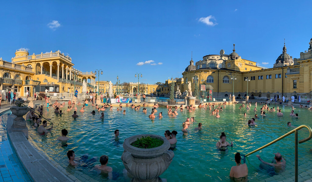 One of the best European cities to visit in November is Budapest - view of the open air thermal baths with lots of people standing in the water and blue sky in background. Copyright@2024 mapandfamily.com 