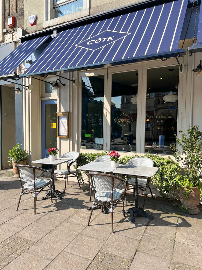 Exterior of Cote Wimbledon with a navy and white striped awning and French-style tables and chairs. Copyright@2024 Nancy Roberts