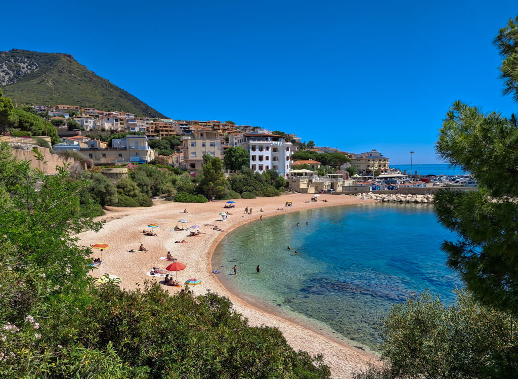 A pretty horseshoe bay of golden sand with low built houses in the distance and greenery in foreground. Copyright@2024 reserved to the photographer vis mapandfamily.com 